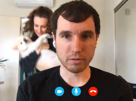 Screen shot of video of man, while a woman dances behind him cradling a dog