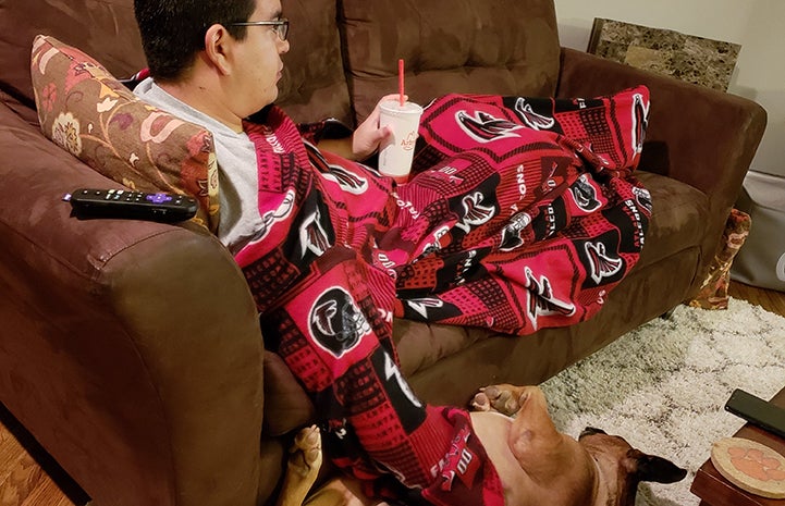 Man lying on a couch under a blanket with Beau the foster dog lying on the floor next to him