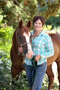Smiling woman wearing a teal plaid shirt next to a brown horse with a white star on his forehead