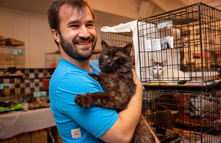 Smiling man holding a dark gray cat at the Best Friends Su-Purrr Adoption event in L.A., with kennels behind them