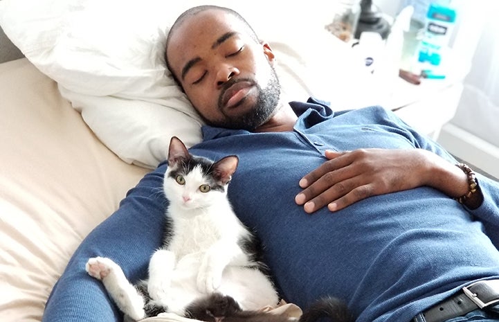 Man sleeping in a bed with a black and white cat cradled up next to him