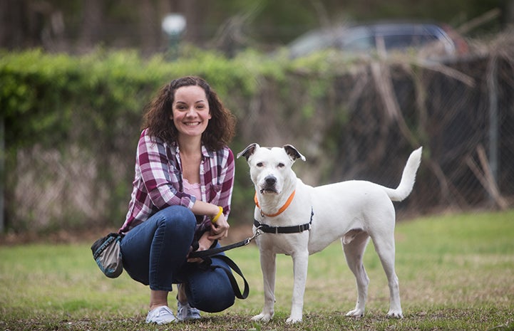 Volunteer Lisa McManus crouching on the ground next to Micah, a white pit bull terrier who is standing and looking at the camera