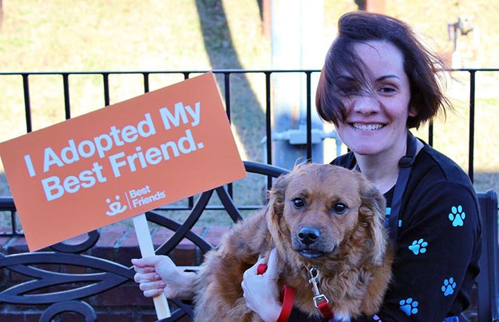 Volunteer Lisa McManus with a brown dog in her lap holding a sign that says, I adopted my best friend