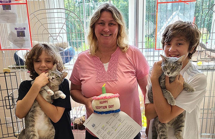 Woman holding a stuffed "Happy Adoption Day" cake and two kids, who are each holding a cat