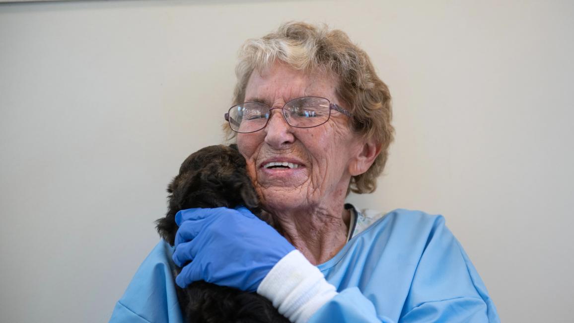 Volunteer Betty Grieb wearing a blue protective gown and gloves, holding a puppy on her shoulder