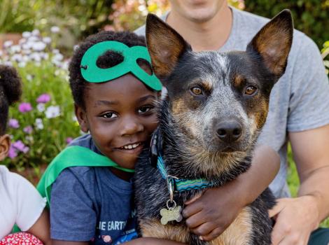 Child wearing a green superhero mask and cape hugging a heeler type dog